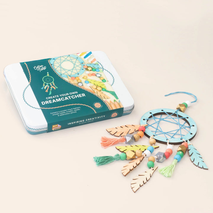 A dreamcatcher craft kit shown with its printed metal tin box and the assembled craft laid out beside it on a plain warm beige background. The kit includes a light blue circular web, wooden feathers in natural tan, colourful beads, and tassels in several pastel colors.