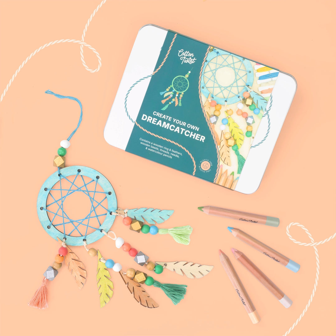 A dreamcatcher craft kit by Cotton Tree shown with its tin packaging on a peach background, alongside a completed light blue dreamcatcher with beads, feathers, and tassels, plus coloured pencils.