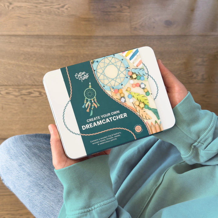 child holding a craft kit packaging with a dreamcatcher design on a wooden floor.