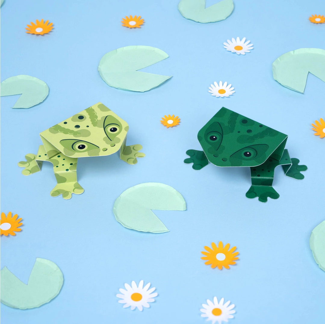 Two green paper frogs on a blue background with flowers and lily pads.