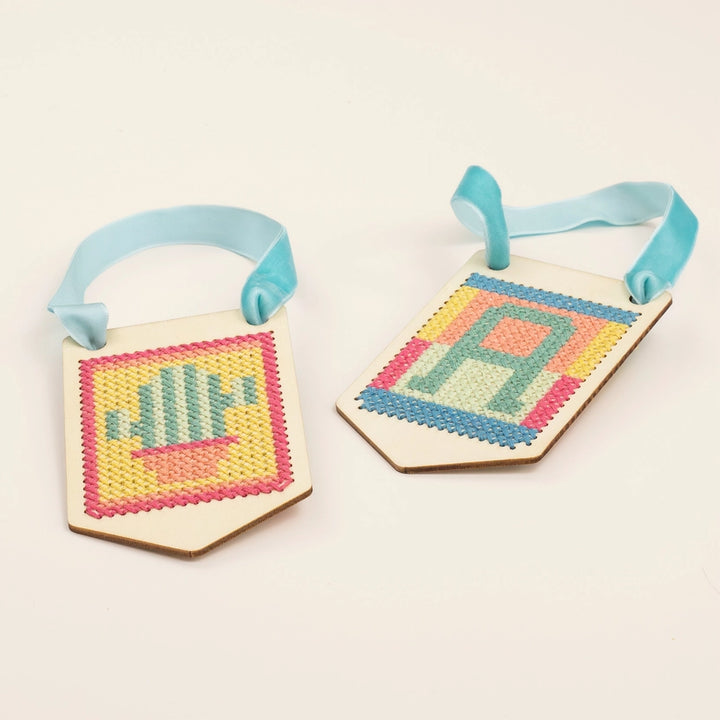 Two cross-stitched tags with colorful patterns and letters on a beige background