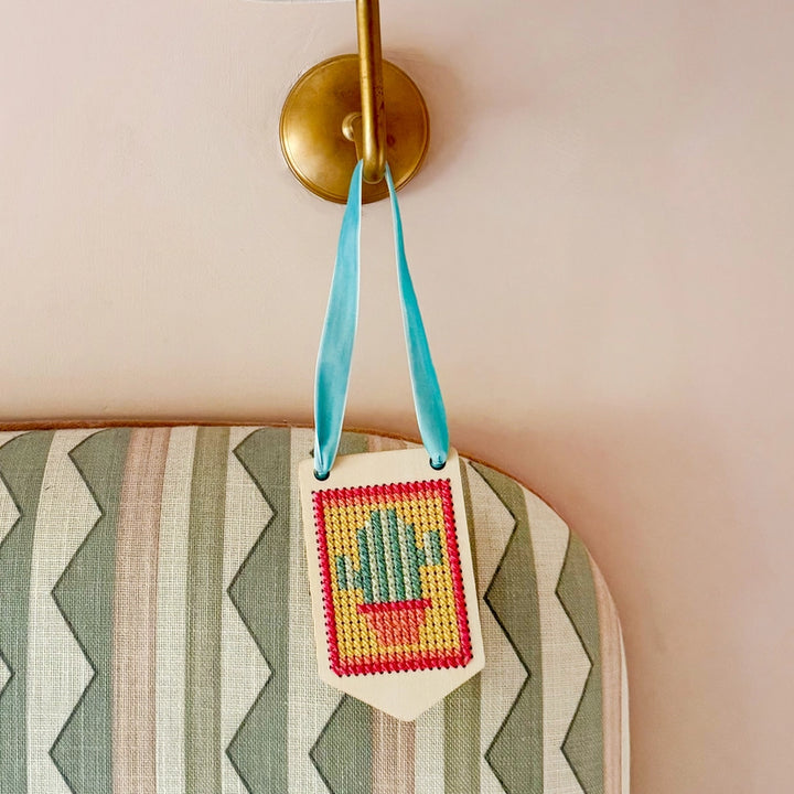 Decorative cross-stitched  banner from the cotton twist make your own stitched banner decoration kit with a cactus design hanging on a wall.