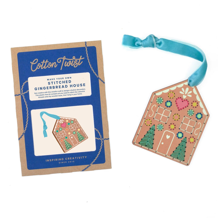 A gingerbread house shaped ornament shown next to its blue and kraft paper packaging card. The ornament features embroidered details in pink, green, yellow, and white on a tan background, with a light blue ribbon loop at the top.