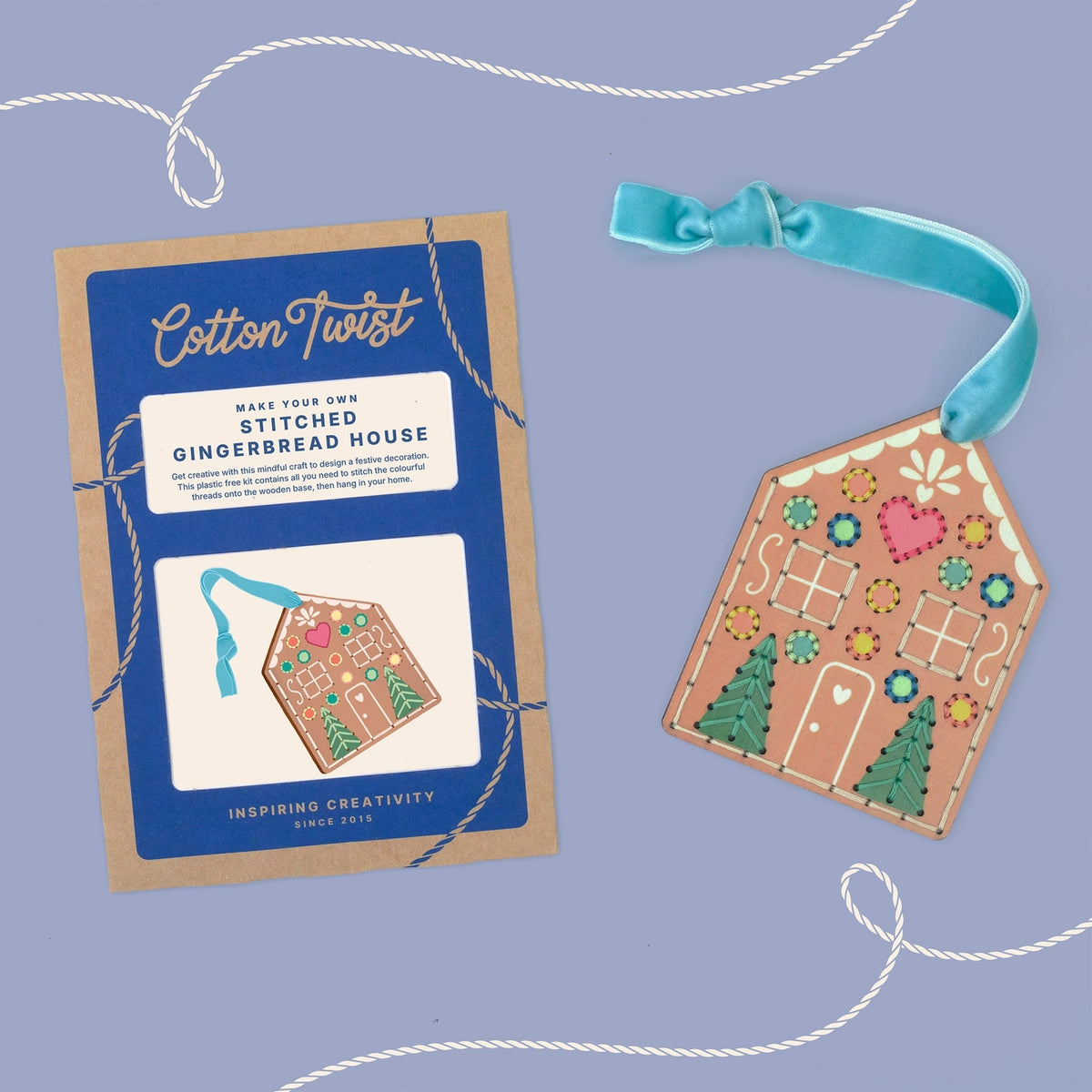 A packaged needlework kit labeled Cotton Twist Stitched Gingerbread House next to the completed embroidered house on a dusty lavender background with decorative white string.