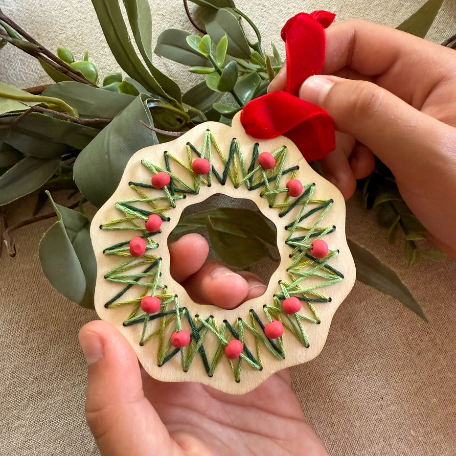 Handmade wooden ornament shaped like a Christmas wreath with greenery and red berries, held by a person against a natural background.