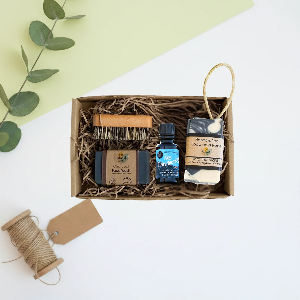 A curated self-care gift set in a brown kraft box filled with shredded paper, featuring a facial brush, two bars of soap, and a small beard oil bottle. The box is photographed from above on a white surface with a light green background panel and decorative twine and eucalyptus props.