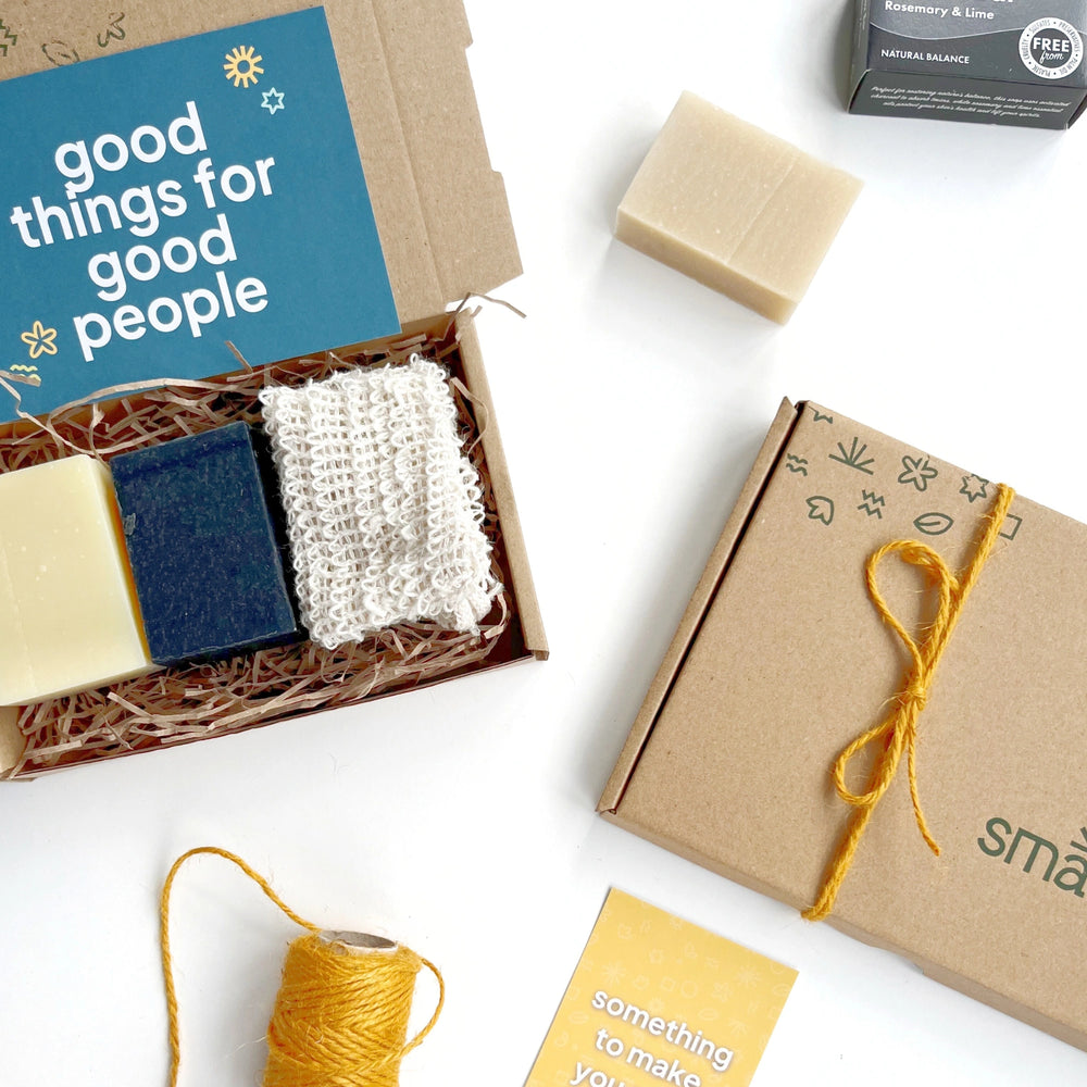 close up of Smallkind mens eco gift set with a black charcoal detox soap, white shaving bar soap and a natural sisal soap saver pouch. The kraft gift box is hand stamped with the smallkind logo and pattern. On top of the box is a green card which reads 'good things for good people'. 