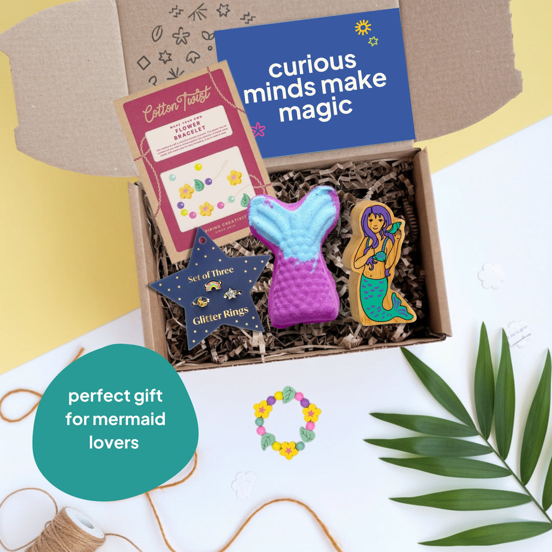 Gift box with mermaid-themed items and 'curious minds make magic' text on a light background.