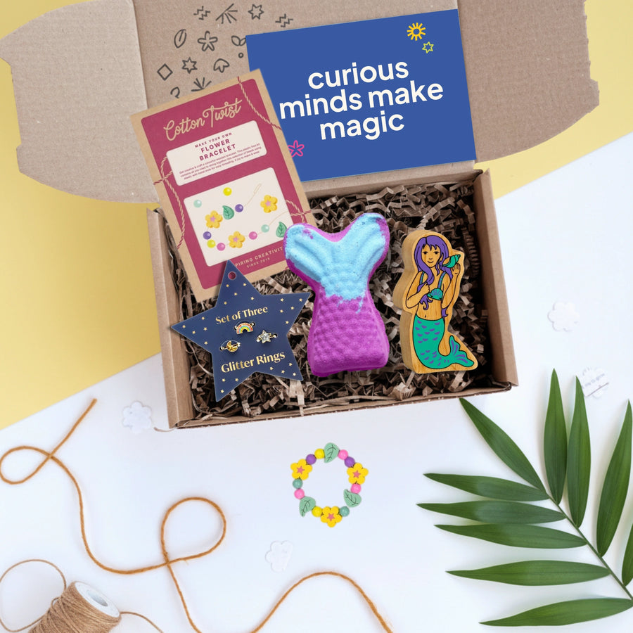 Gift box filled with mermaid themed toys, crafts and a bath bomb with 'curious minds make magic' card on a yellow and white background