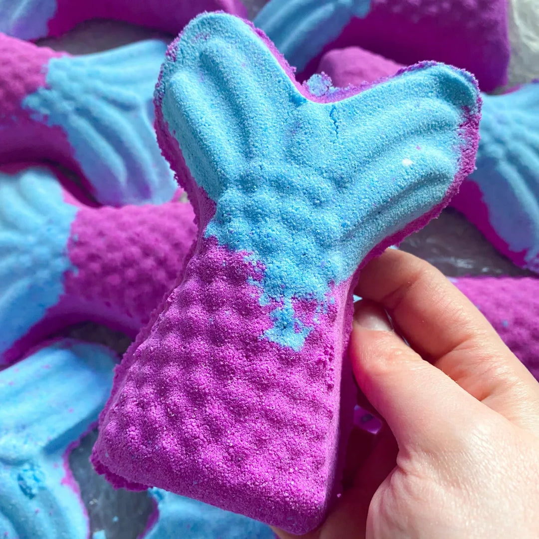 Purple and blue mermaid tail bath bomb held by a hand with more bath bombs in the background.
