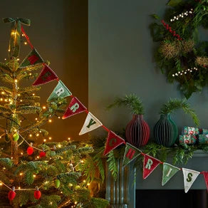 A festive Christmas garland banner spelling out the months of the year in red, green, and white triangular flags, hung in a decorated holiday room next to a lit Christmas tree and mantel.