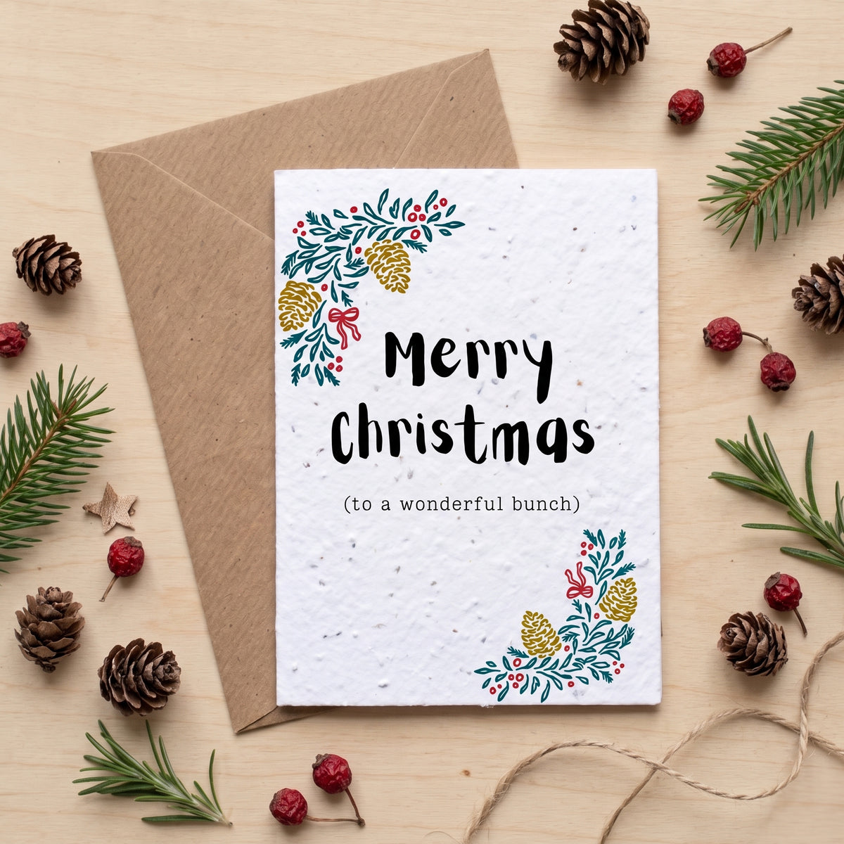 A square white Christmas greeting card with floral greenery and red berry illustrations in the corners, reading 'Merry Christmas (to a wonderful bunch)'. It is shown with a brown kraft envelope on a light wood surface, surrounded by pine branches, pinecones, and red berries.
