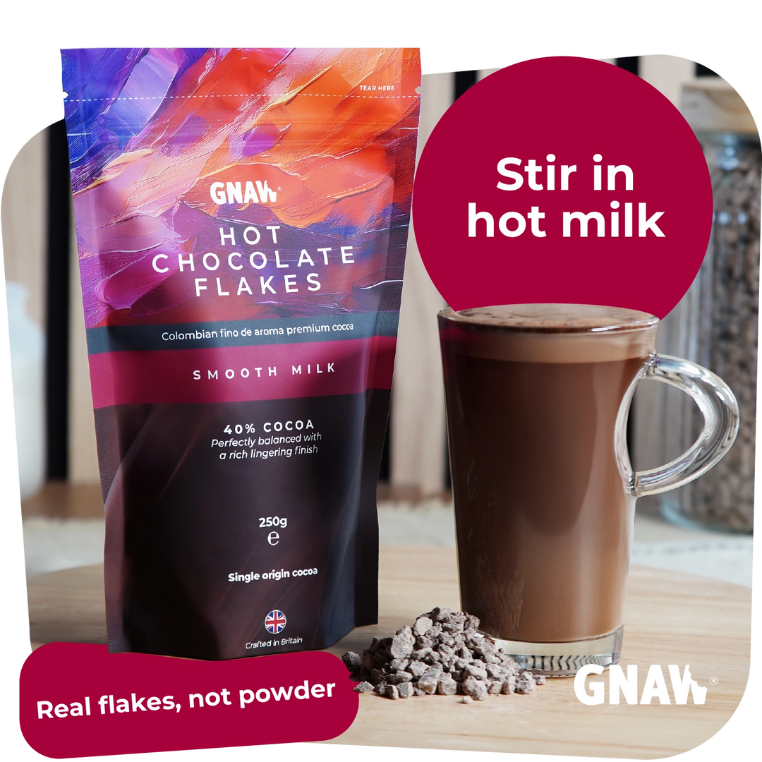 A bag of GNAW Hot Chocolate Flakes in smooth milk flavor stands next to a glass of chocolate drink with a pile of chocolate flakes on a tabletop. A pink circle says &