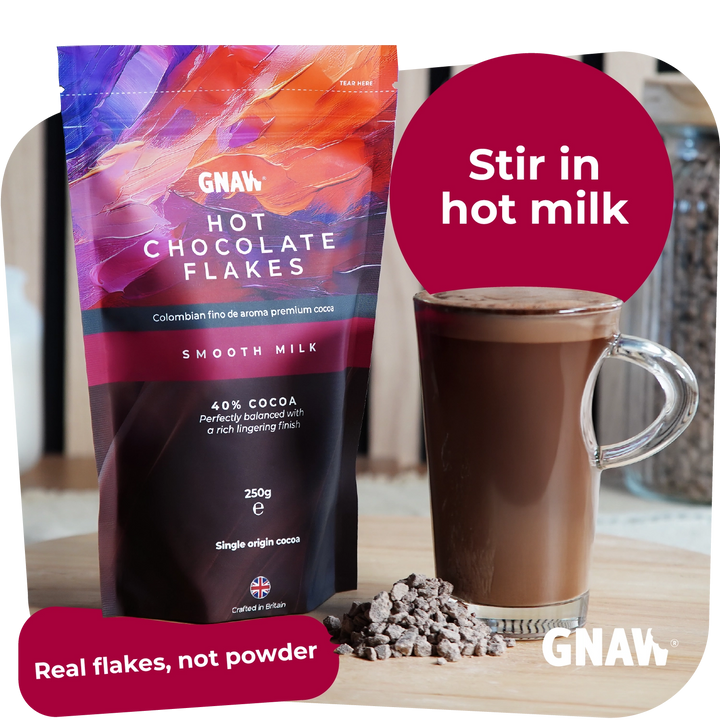 A bag of GNAW Hot Chocolate Flakes in smooth milk flavor stands next to a glass of chocolate drink with a pile of chocolate flakes on a tabletop. A pink circle says &