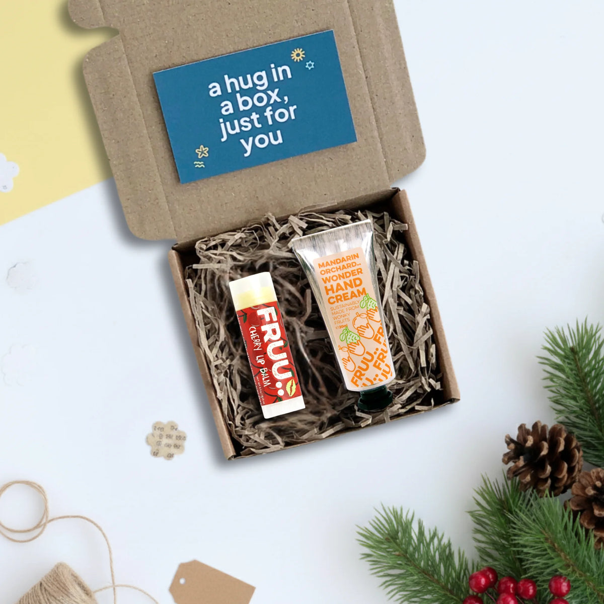 A curated gift box set featuring a tube of FRUU lip balm and a tube of Mandarin Hand Cream nestled in shredded paper inside a brown kraft box, with a blue card reading 'a hug in a box, just for you' on the lid, styled with pine branches and pinecones on a white surface.