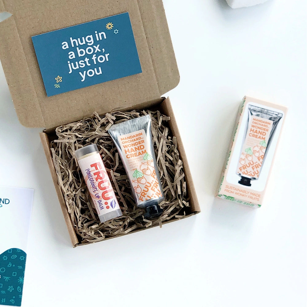 a mandarin orchard hand cream and pomegranate lip balm, both made by Fruu cosmetics, inside a krfat gift box filled with shredded paper. The gift card is dark green and has a message 'a hug in a box just for you' in bold white text. 