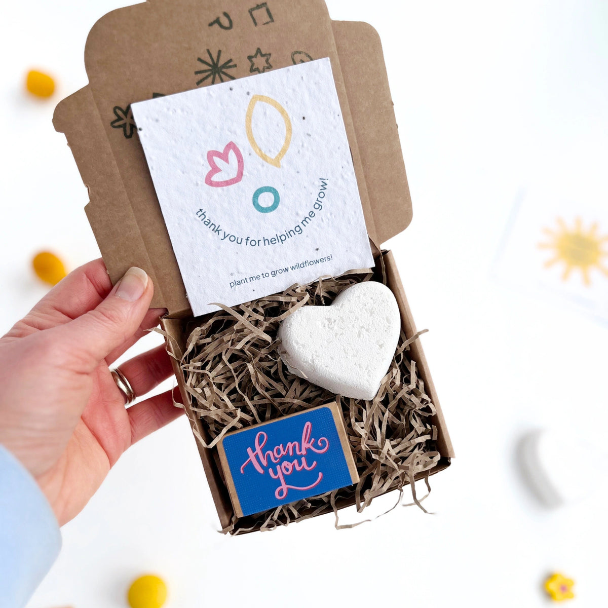 womans hand holding a kraft gift box with heart shaped bath bomb, blue box of thank you seedballs and a seedpaper card which says 'thank you for helping me grow'