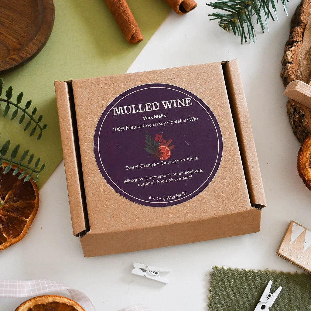 A kraft paper box of 'Mulled Wine' wax melts by The Natural Spa is displayed in a styled flat-lay arrangement with holiday-themed props like pine needles, dried orange slices, and cinnamon sticks on a white and green background.