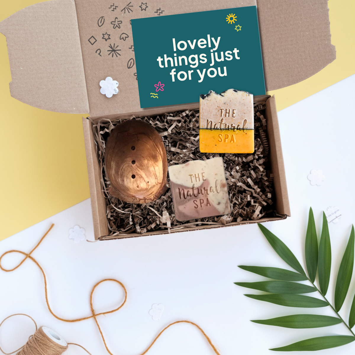 An open gift box containing an olive wood soap dish and two handmade soap bars labeled 'The Natural Spa' on shredded paper, with a teal card reading 'lovely things just for you' inside a styled flat-lay scene.