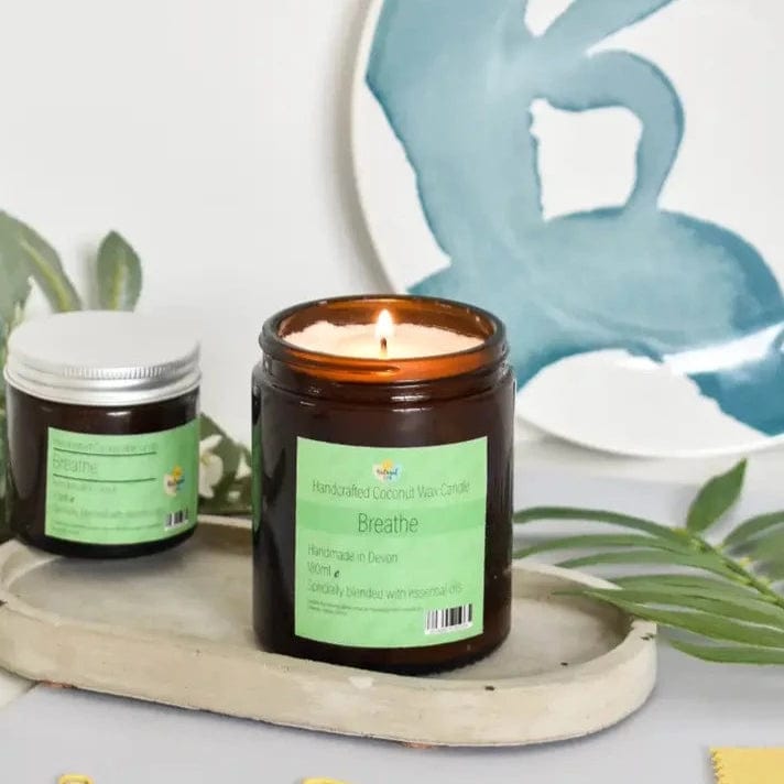 A lit coconut wax candle in an amber glass jar with a green label, set on a wooden tray with a larger jar behind it, against a blue and white background.