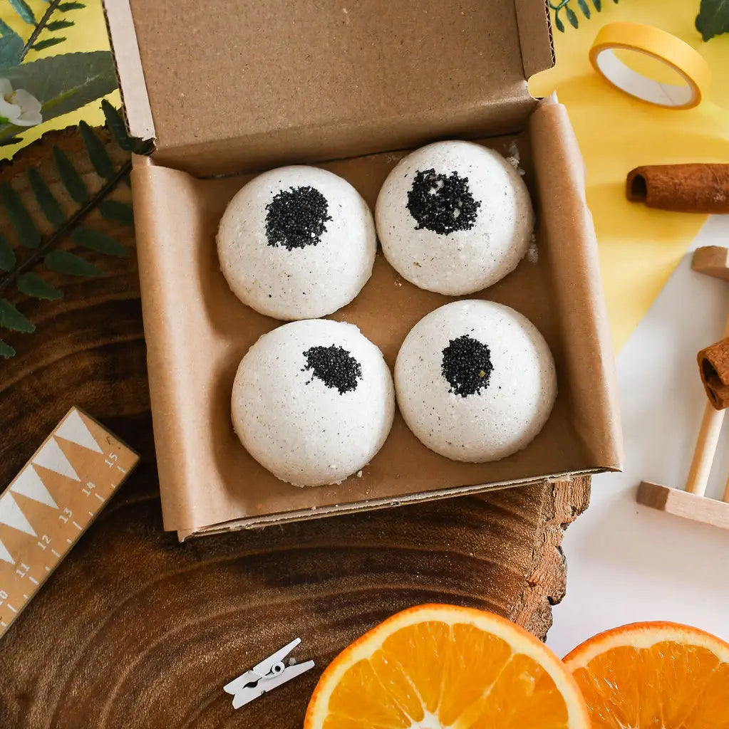 Four aromatherapy shower steamers topped with cardamom seeds in a cardboard box, surrounded by oranges and a wooden surface.