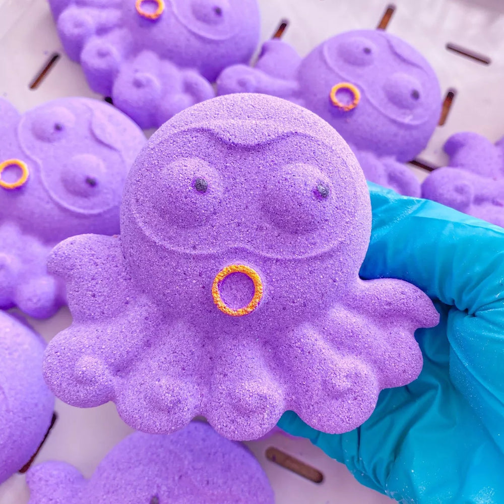 Purple octopus-shaped bath bomb with a yellow ring held by a blue gloved hand.