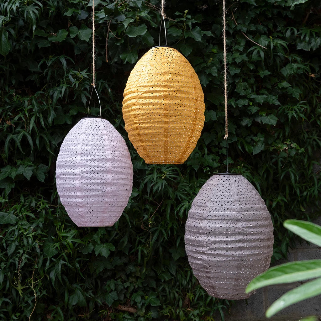 Three hanging paper lanterns in yellow, white, and gray against a green leafy background.