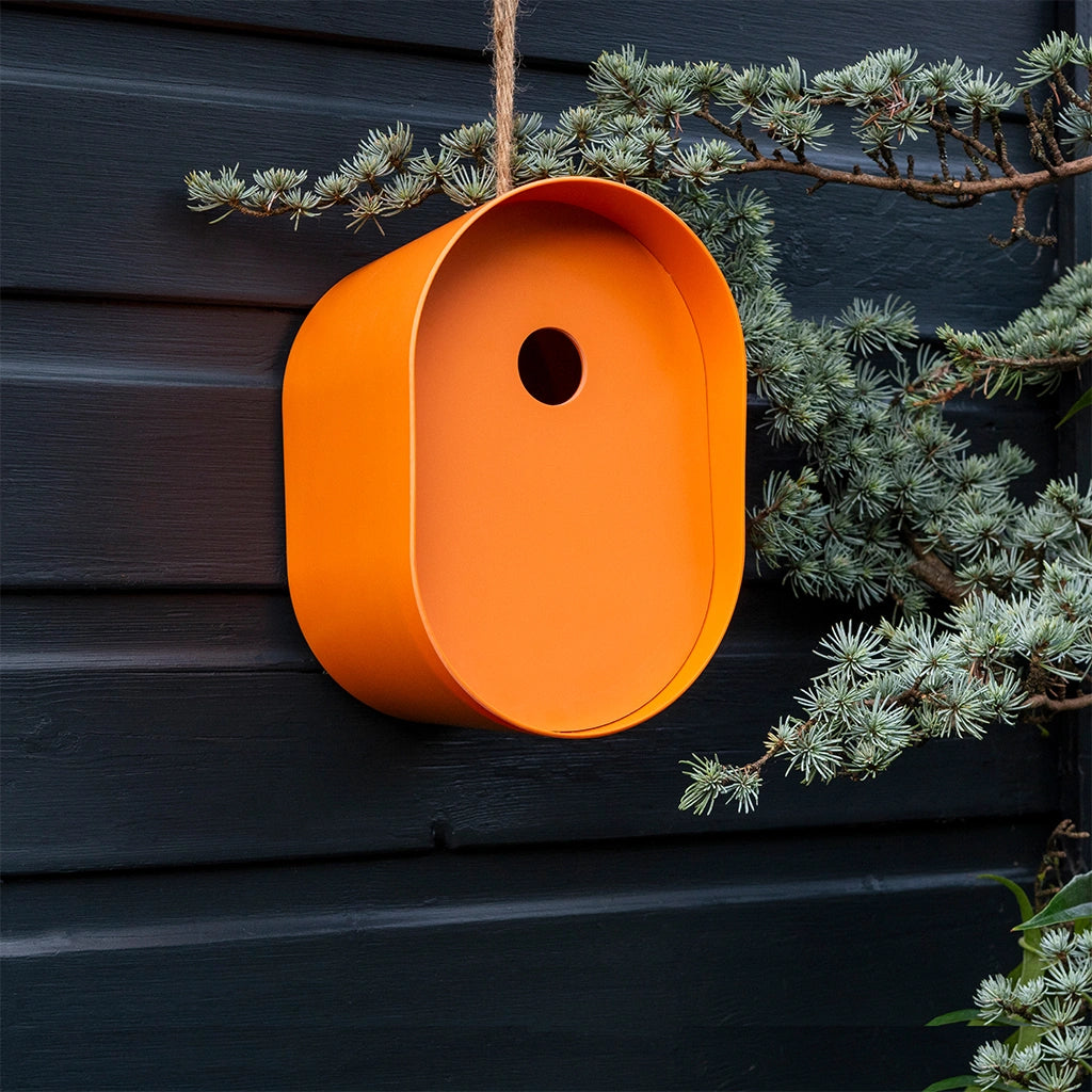 contemporary Rex London Oval Bamboo Bird House in a striking orange with rope hanging loop. Hung against a grey fence
