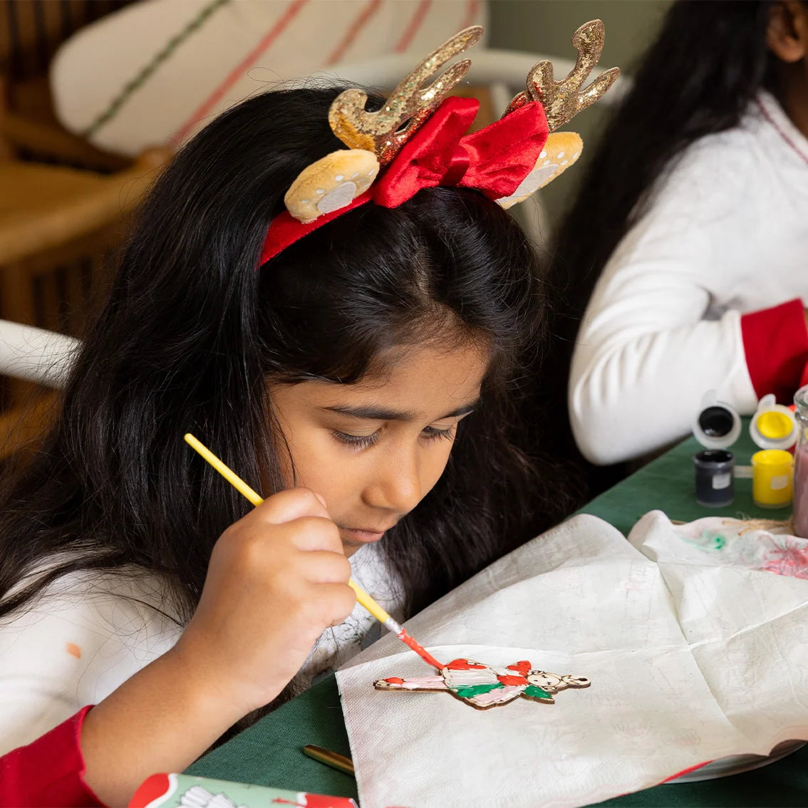 Child wearing reindeer antlers painting a gingerbread cookie with a brush.