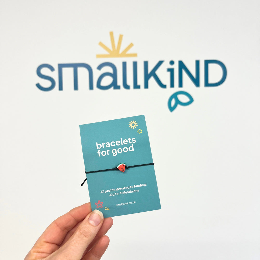 white womans hand holding a watermelon friendship bracelet on a green card . The card has white text which reads 'bracelets for good'. The smallkind logo is on the wall in the background.