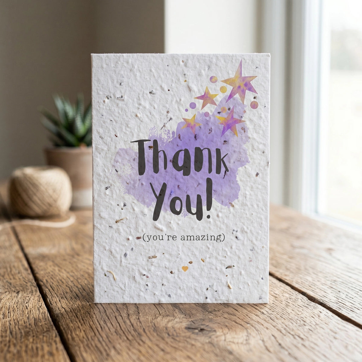 Greeting card with 'Thank You! (you're amazing)' text and purple watercolor design on a wooden surface.
