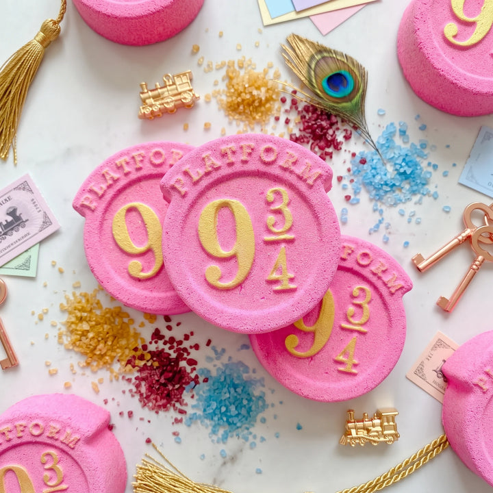 several pink platform 9 inspired bath bomb with gold details. The background includes magical decorative elements such as peacock feathers and gold keys. 