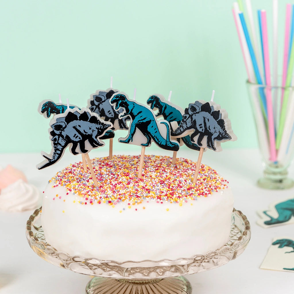 six dinosaur shaped birthday candles in a white cake with sprinkles 