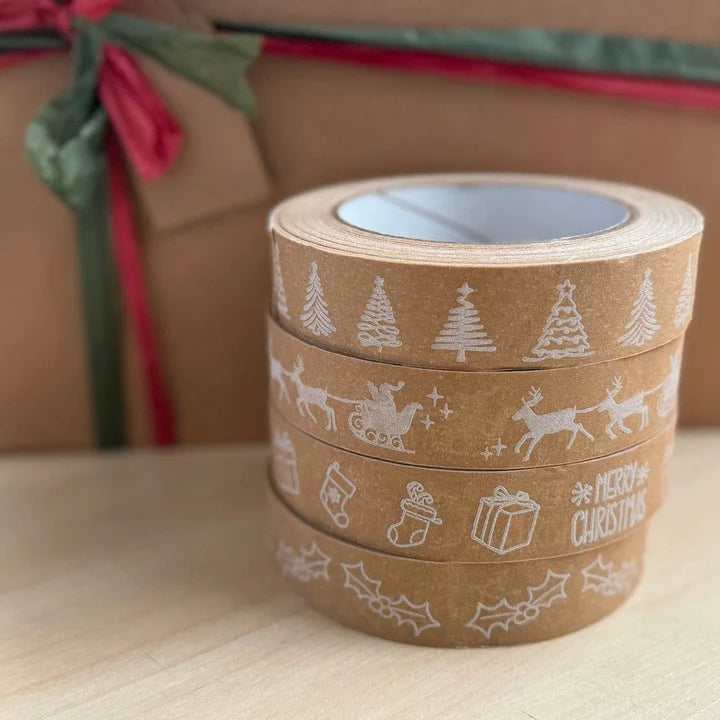 stack of four rolls of Christmas-themed decorative paper tape printed with white festive prints. The tapes are on a wooden surface with a gift box in the background.