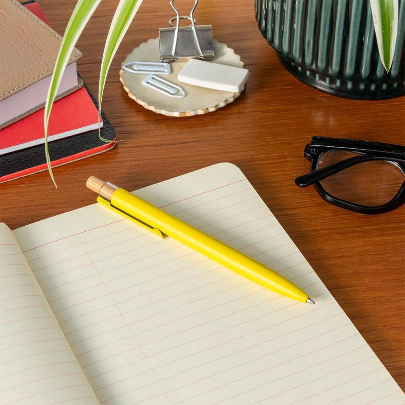 Open notebook with a yellow pen on a wooden desk, with books and a plant in the background.