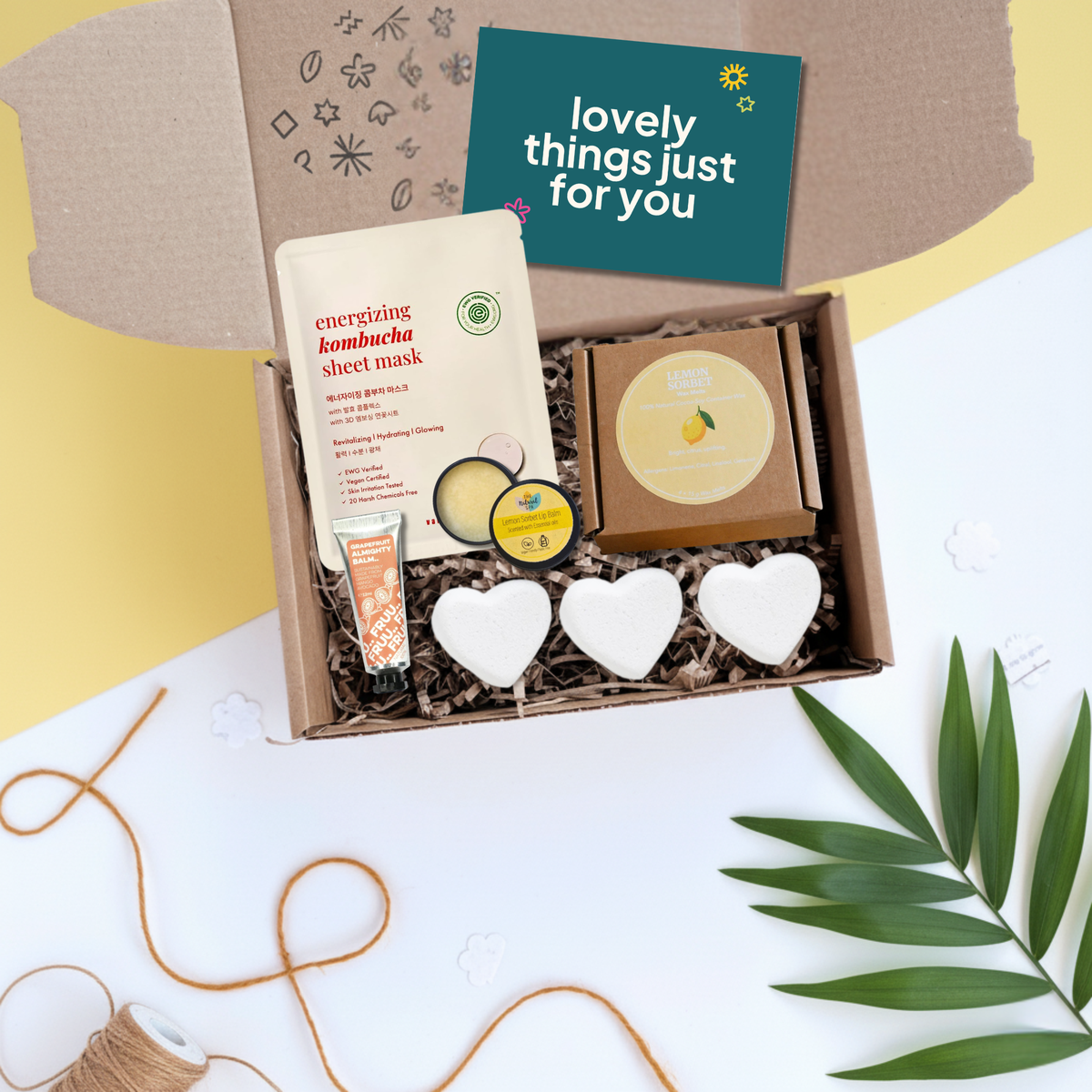 A curated gift box set containing a sheet mask, a tube of grapefruit almighty balm, lemon wax melts, and three white heart-shaped bath bombs, arranged in a brown cardboard box with shredded paper filler. The box lid is open, showing a teal card that reads 'lovely things just for you'