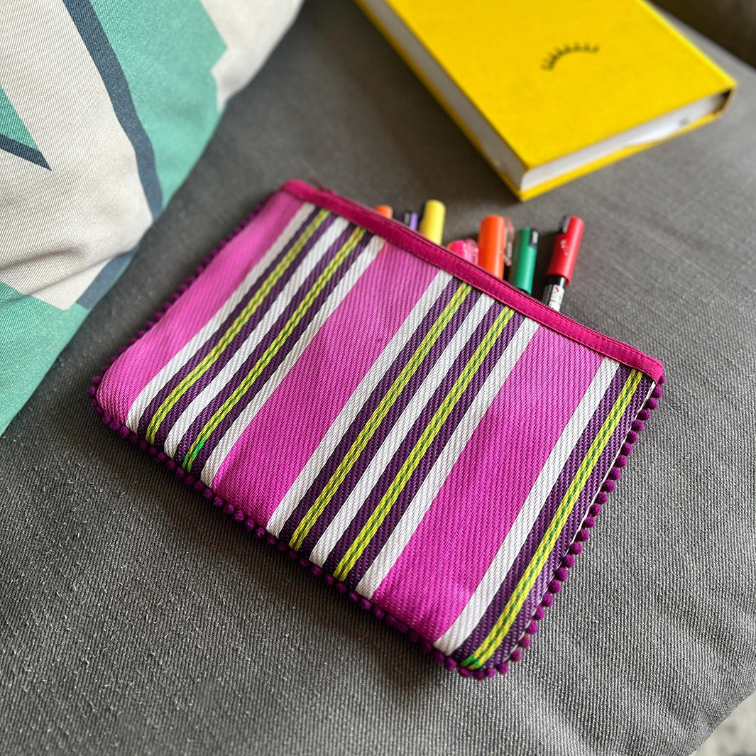 Respiin Recycled Woven Pom Pom Pouch in bright fuchsia pink with purple, yellow and white stripes and a pink pom pom trim. The pouch is on a sofa with stationery and pens inside.