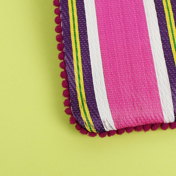 close up of a corder of a Respiin Recycled Woven Pom Pom Pouch in bright fuchsia pink with purple, yellow and white stripes and a pink pom pom trim. 