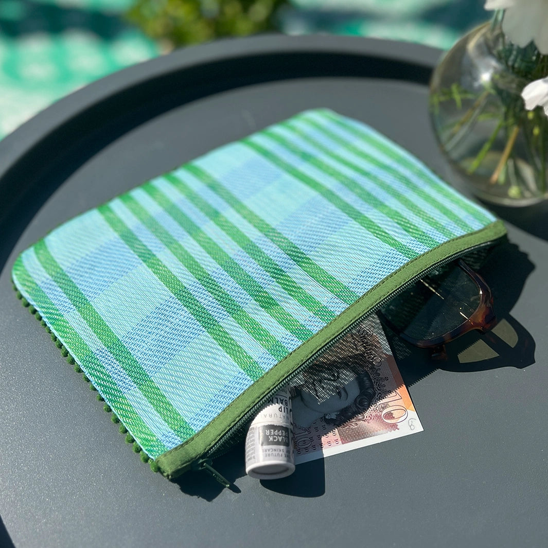  Respiin Recycled Woven Pom Pom Pouch in a blue and green checked design with a green pom pom trim. The pouch is open with sunglasses, money and lip balm inside.