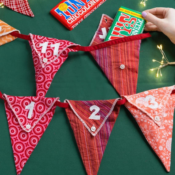 Red and pink patterned reusable Christmas advent calendar bunting with numbers on a green surface with chocolate bars and fairy lights.
