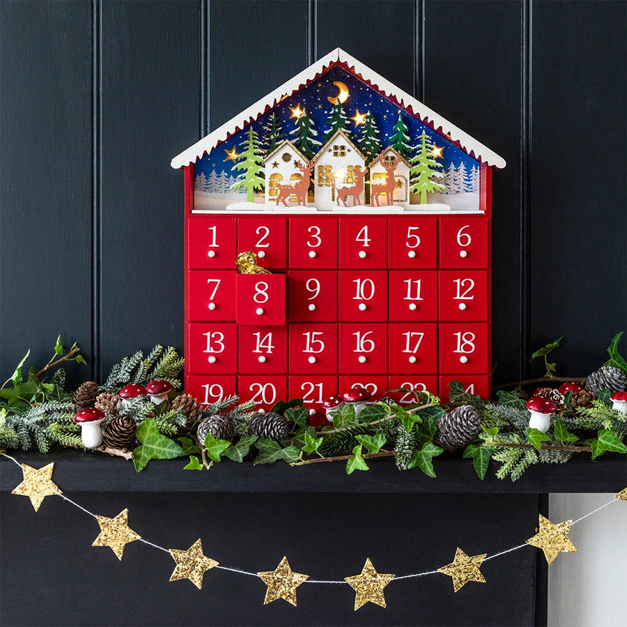 Red advent calendar with Christmas scene on top, decorated with greenery and stars on a dark background.