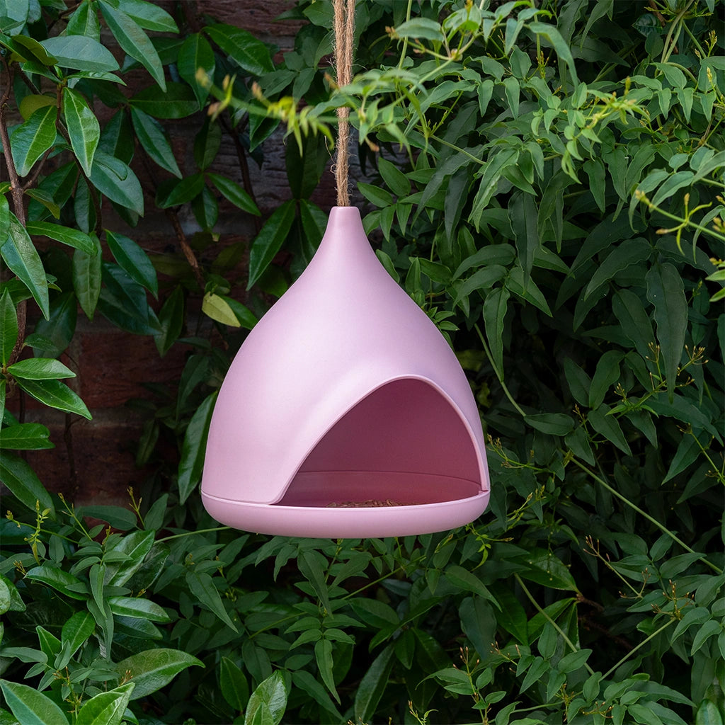pastel pink bamboo bird feeder with a hanging rope. the feeder hangs from a leafy bush outdoors. 