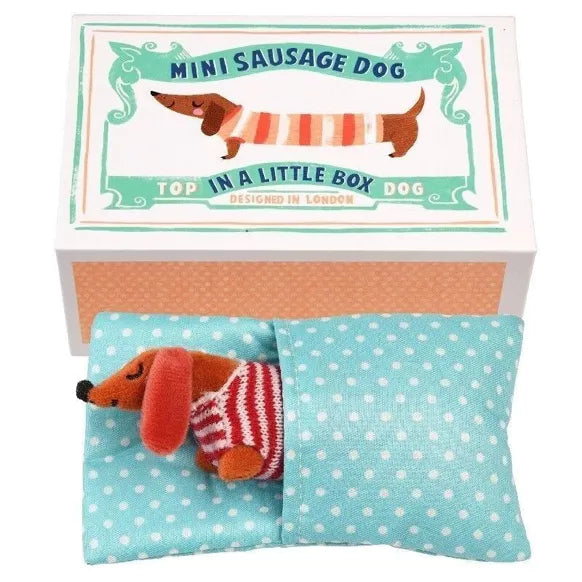 A plush toy of a sausage dog in a striped jumper, presented in a box with a blue polka dog bed. for age 3+