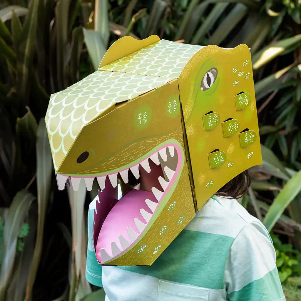 Child wearing a green and yellow 3D dinosaur head mask, showing the assembled kit's open mouth with white teeth and pink tongue, against a background of lush green plants