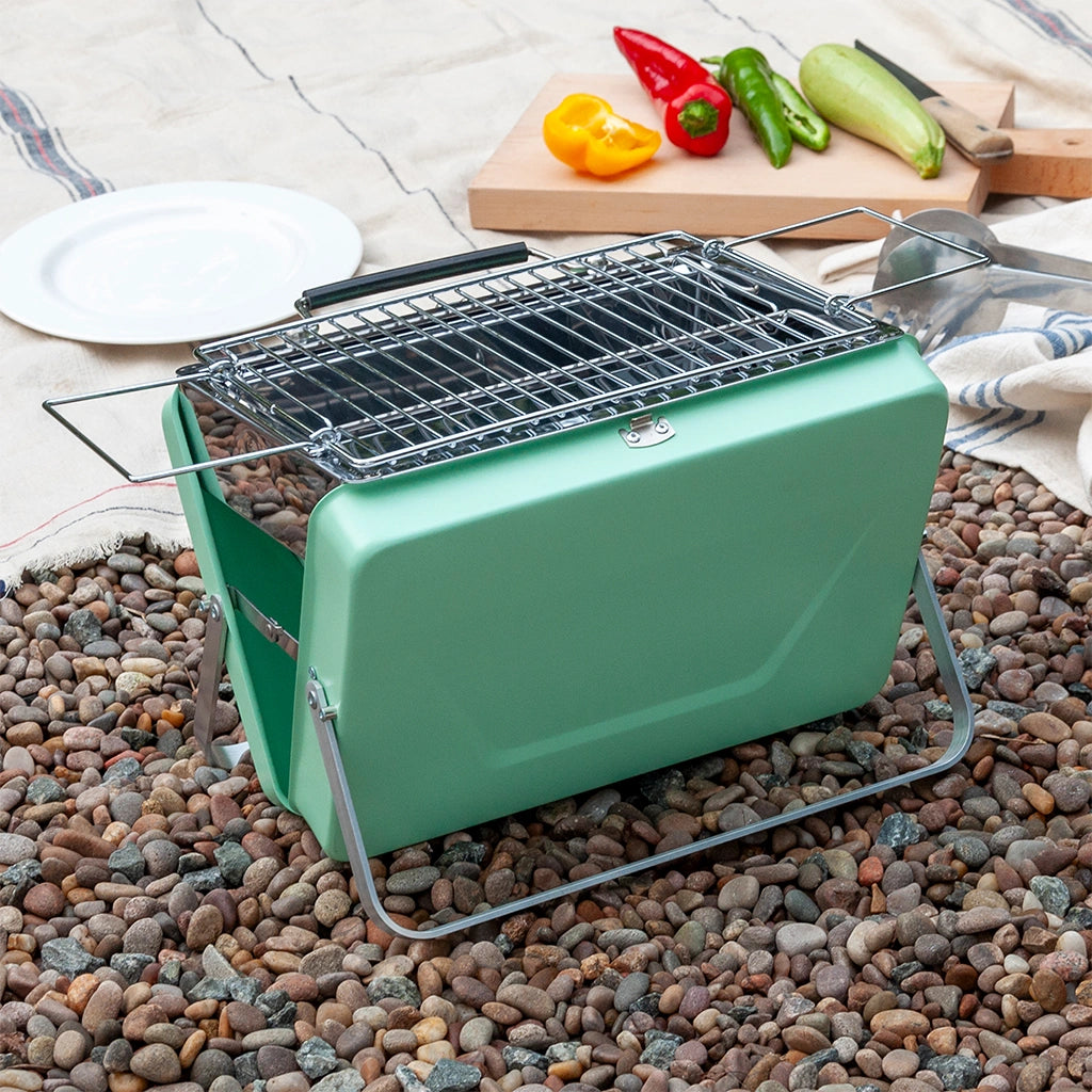 Rex London Portable Suitcase BBQ in a stylish pistachio green. The barbecue is on gravel with a picnic blanket in the background.