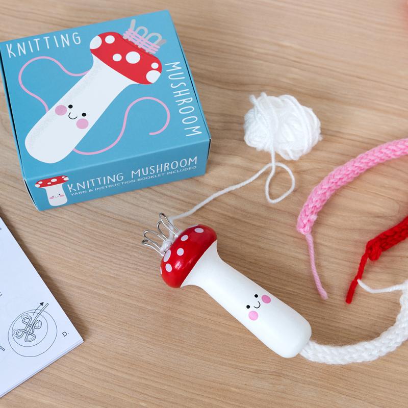 Mushroom shaped spool knitter for kids with a cute painted toadstool design. The knitting mushroom is on a wooden table with red, pink and white wool and a blue gift box.