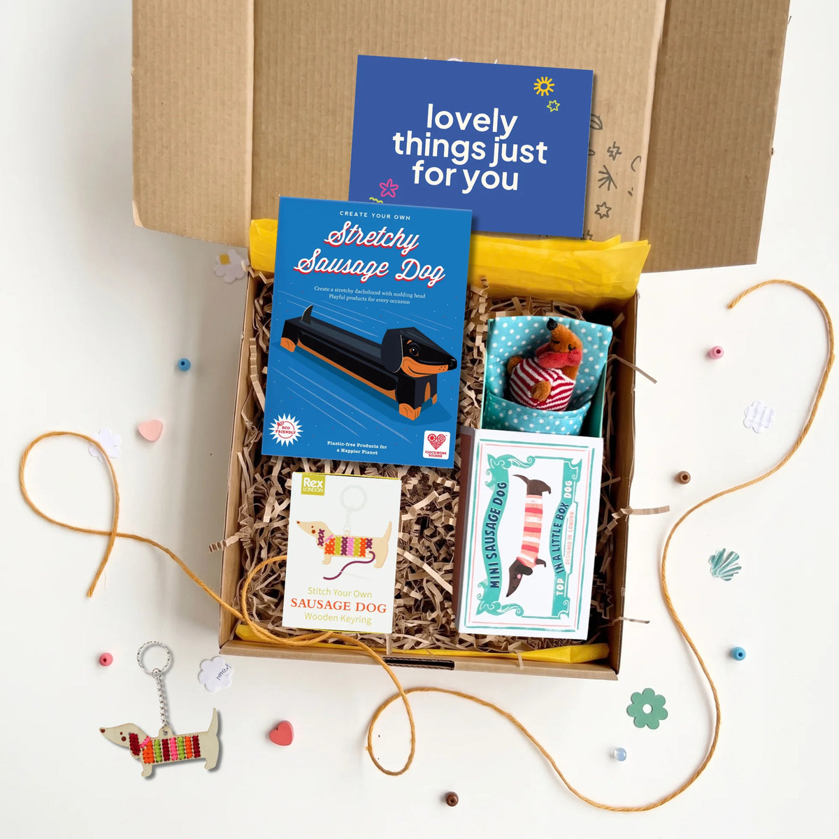 A curated dog-themed gift box filled with shredded paper and yellow tissue paper, containing a blue 'Stretchy Sausage Dog' craft kit, a small plush dog, a card, and a small keyring craft kit An open brown cardboard box sits behind the gift box on a white surface with scattered decorative paper hearts, string, and dog-shaped charms.