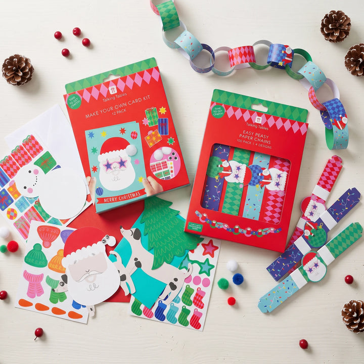 Two Christmas-themed card-making kits in red packaging are displayed on a light wood surface with colorful paper chains, stickers, and small pinecones around them.