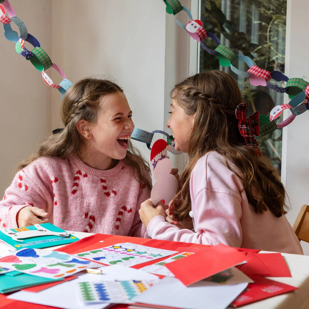 Two young girls laughing and blowing into a small pink and white toy whistle, sitting at a table covered with colorful paper crafts and cards. A paper chain hangs in the background near a window.