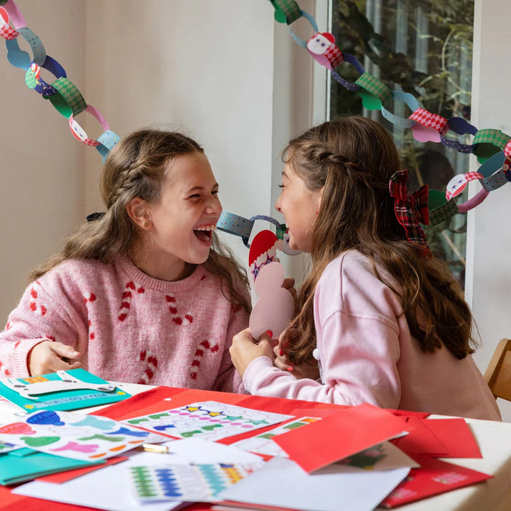 Two young girls laughing and blowing into a small pink and white toy whistle, sitting at a table covered with colorful paper crafts and cards. A paper chain hangs in the background near a window.